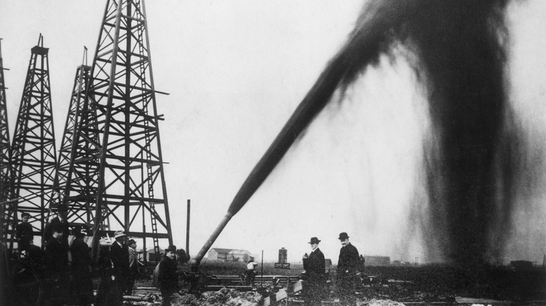 An early gusher blowing over wooden derricks, with men in bowler hats standing at the wellhead.